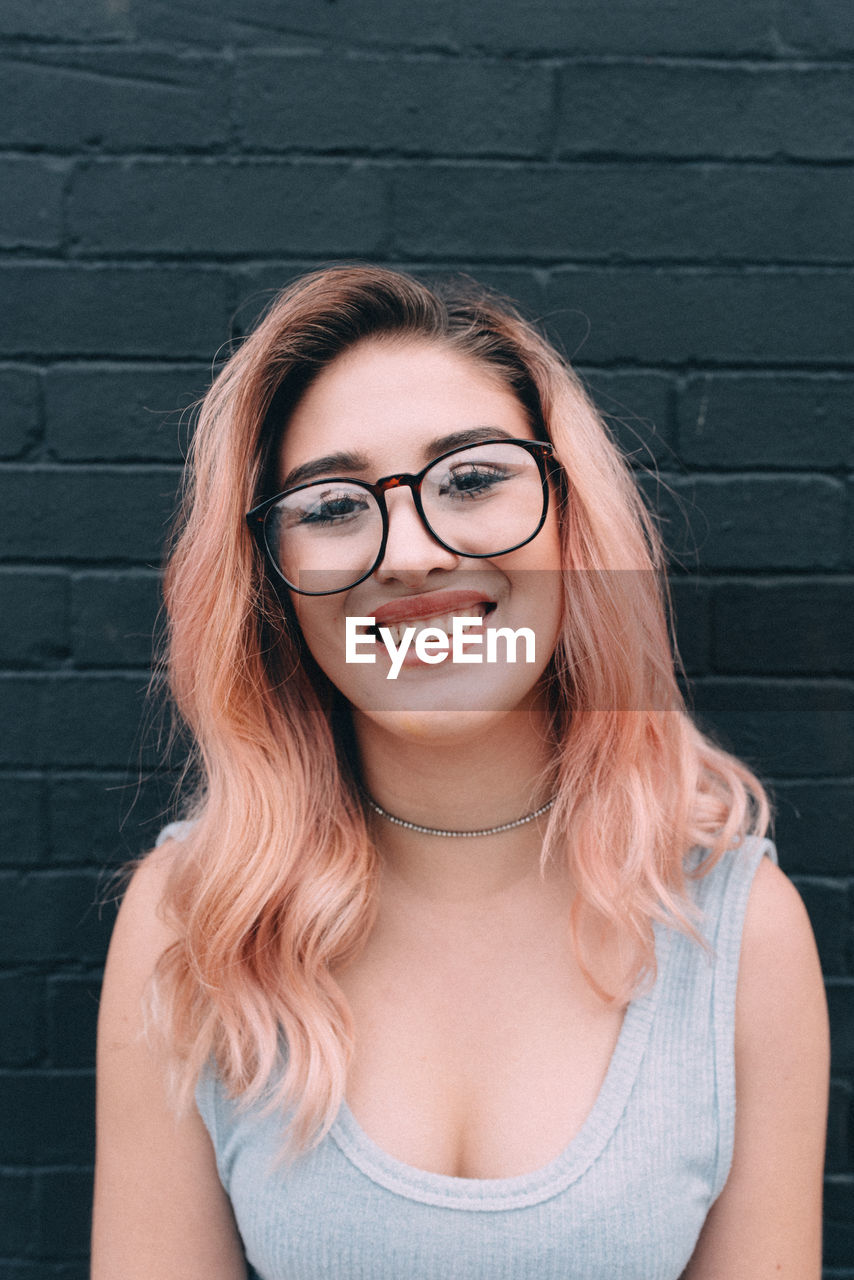 Portrait of cheerful young woman wearing eyeglasses against black brick wall