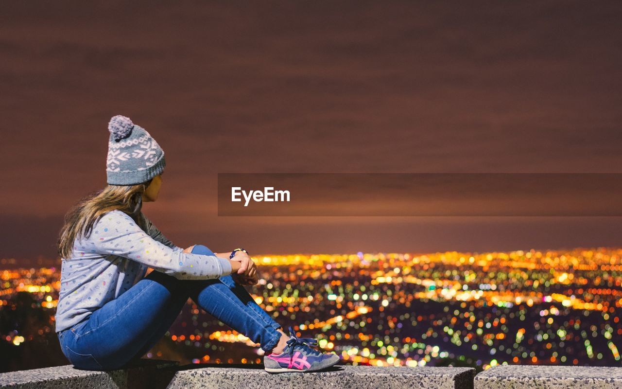 Full length of woman sitting on building terrace against illuminated city at sky high mount dandenong
