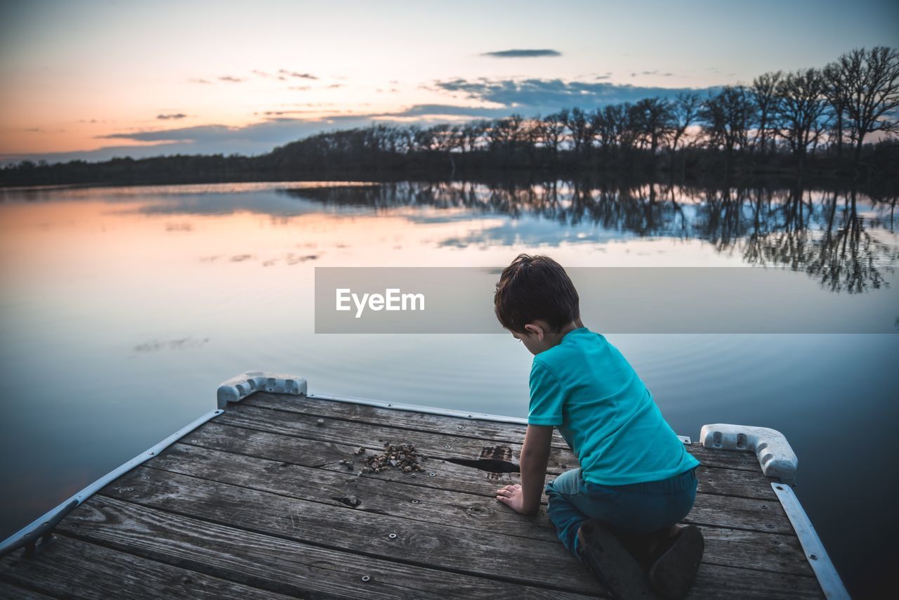 Boy on jetty over lake against sky during sunset