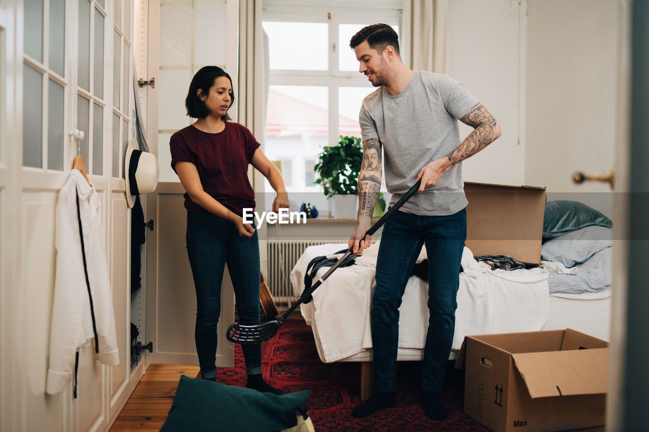 Woman gesturing while man holding golf club by bed in bedroom during relocation