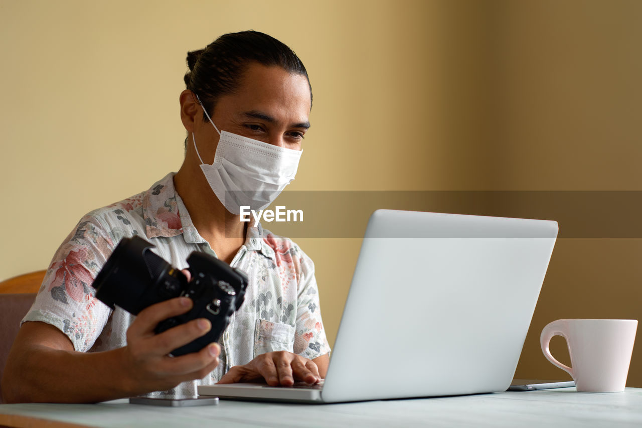 Man wearing mask holding camera while using laptop