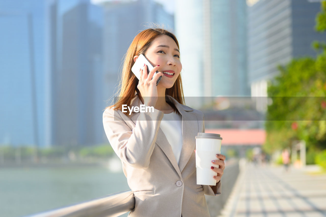 Businesswoman holding coffee talking over smart phone while standing outdoors