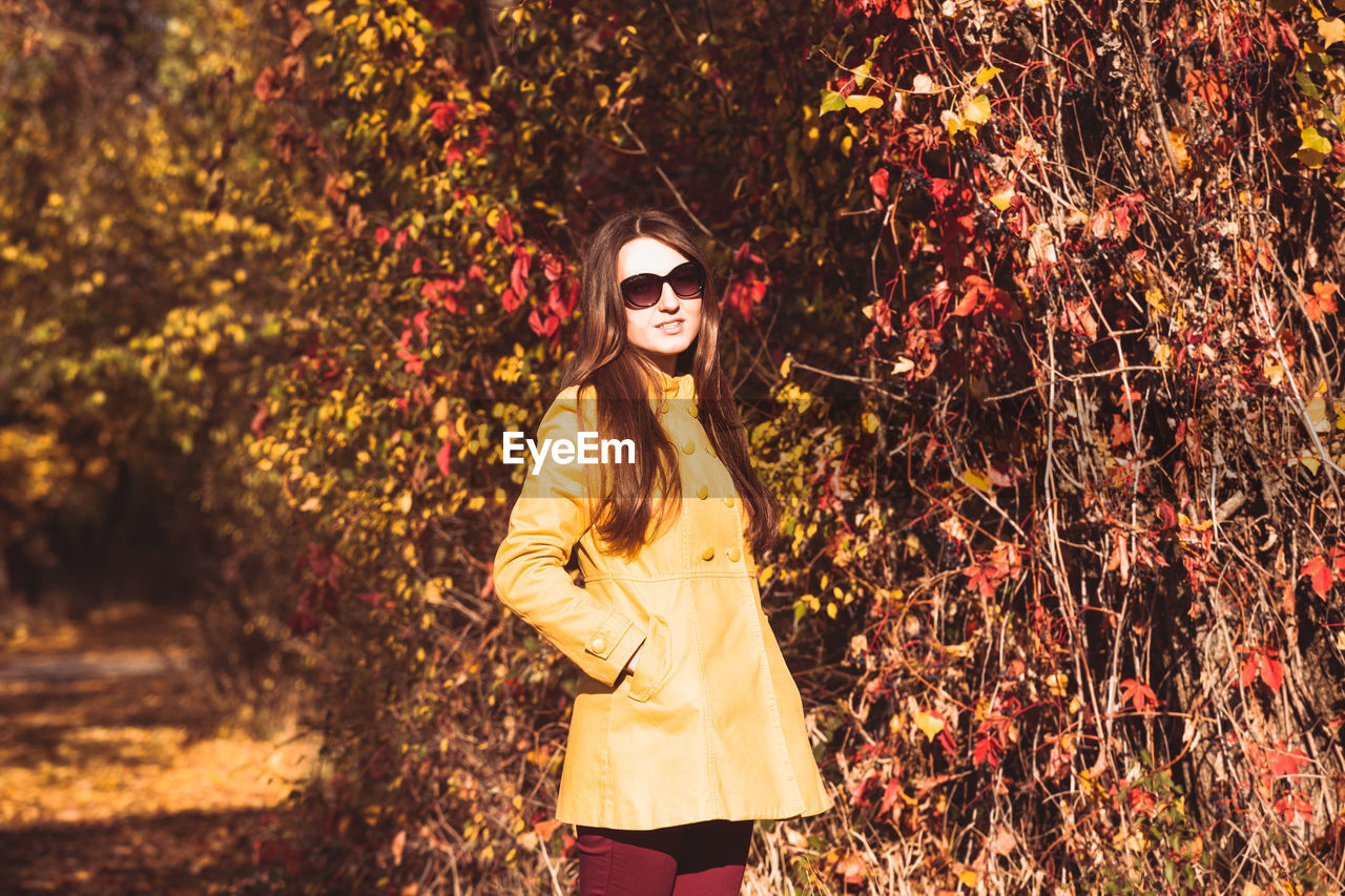 PORTRAIT OF YOUNG WOMAN WEARING SUNGLASSES STANDING BY TREE
