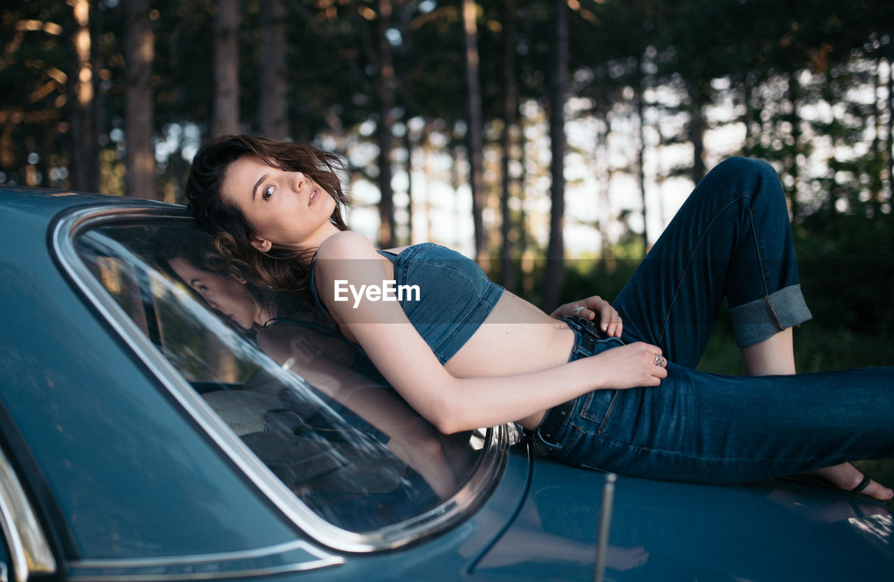 Hipster woman lying on vintage car in the forest.