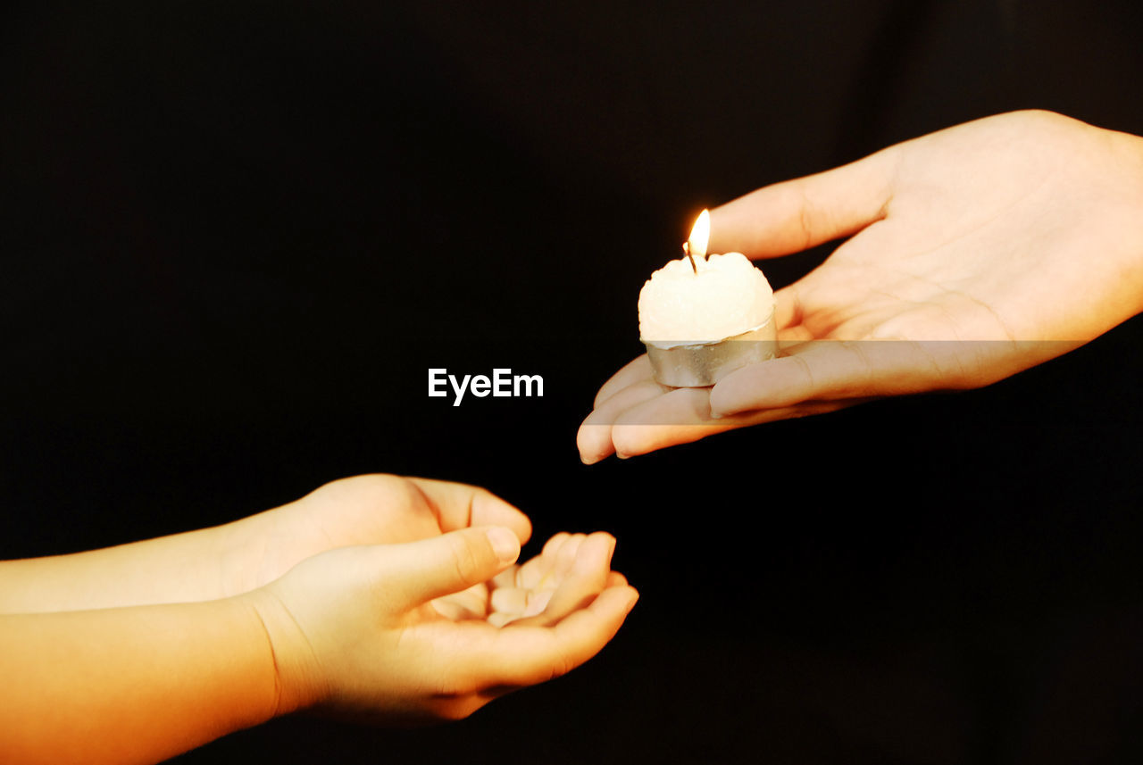 Cropped hand giving lit tea light to child against black background