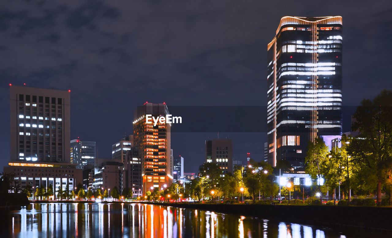 ILLUMINATED BUILDINGS BY RIVER AGAINST SKY