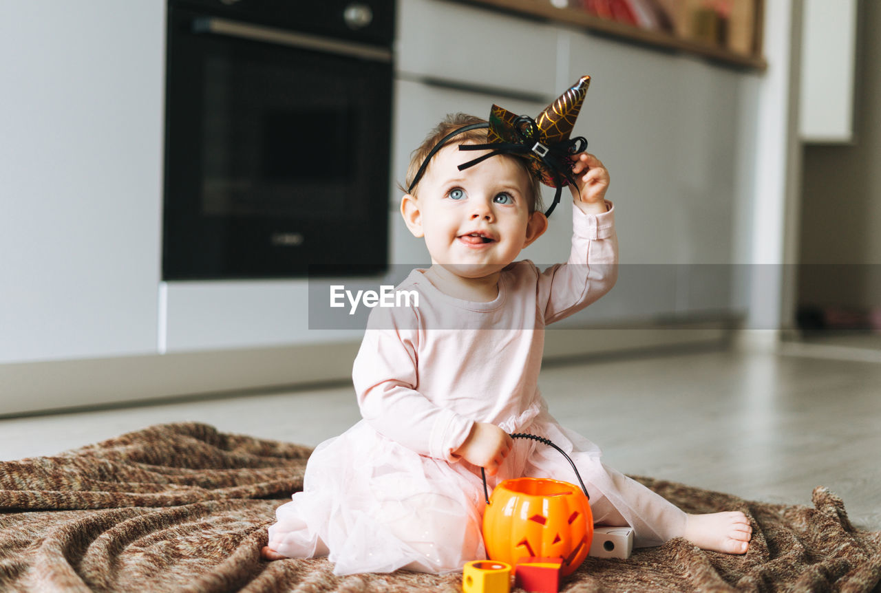 Cute little baby girl in witch's hat and pumpkin lantern in kitchen at home, halloween time