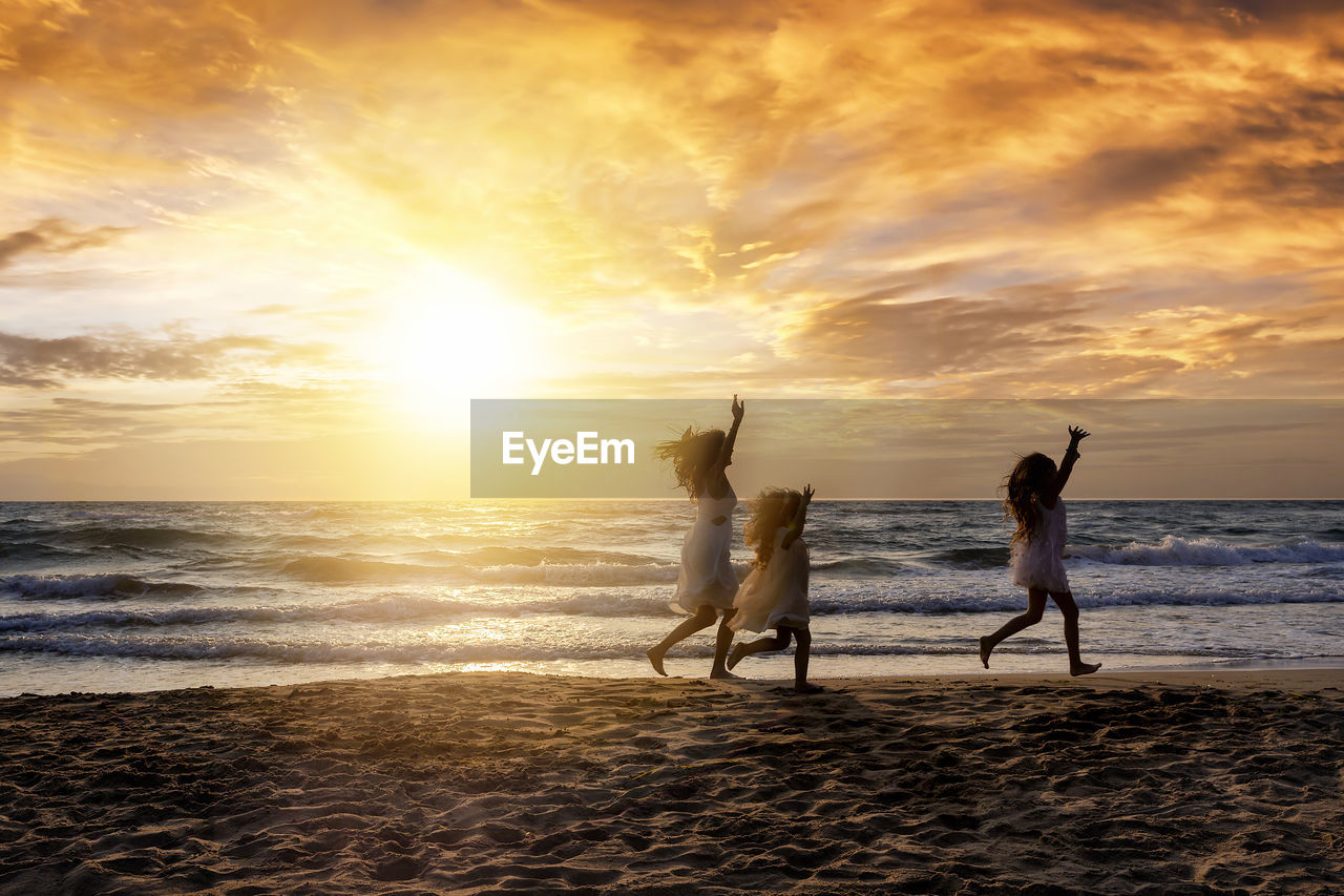 Side view of mother and daughters with arms raised running at beach against orange sky