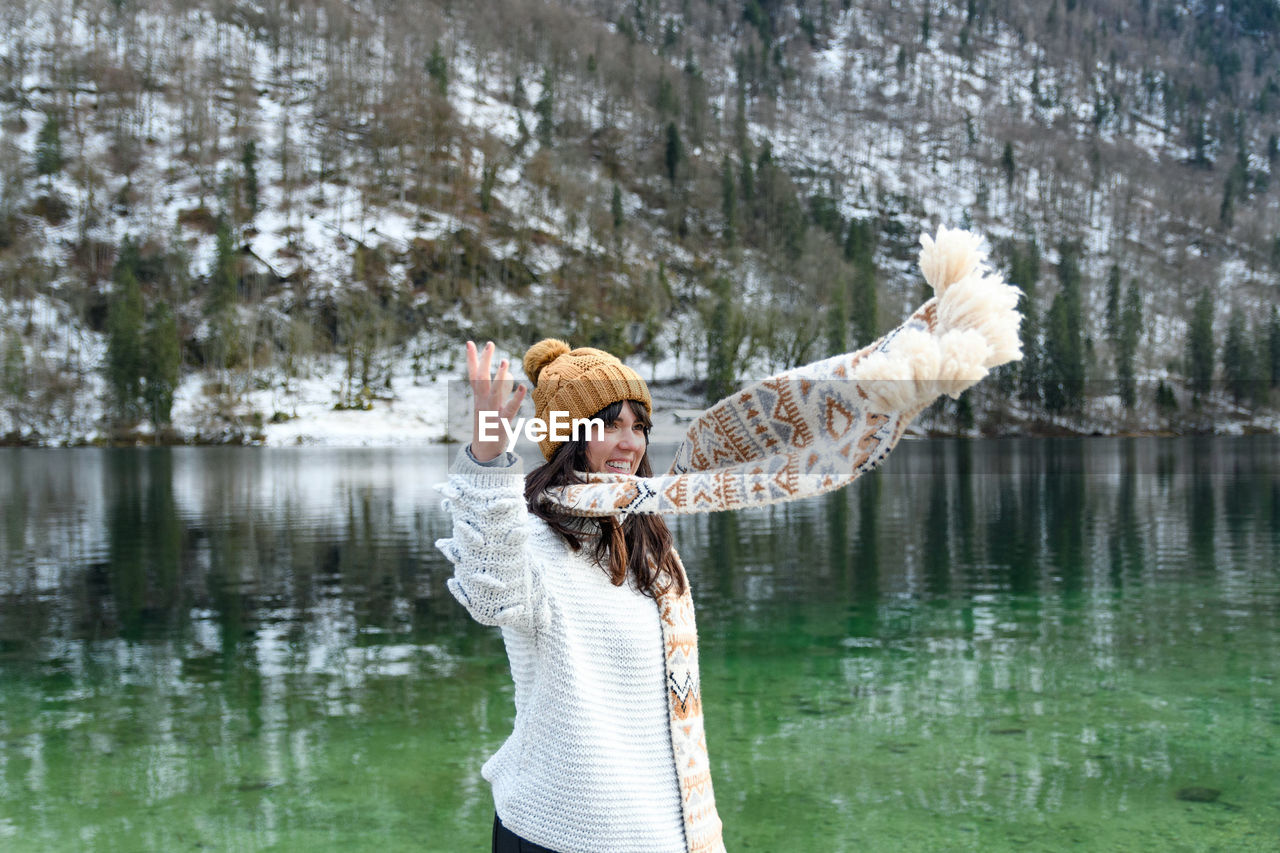 Young woman in winter sweater smiling, standing by a lake.