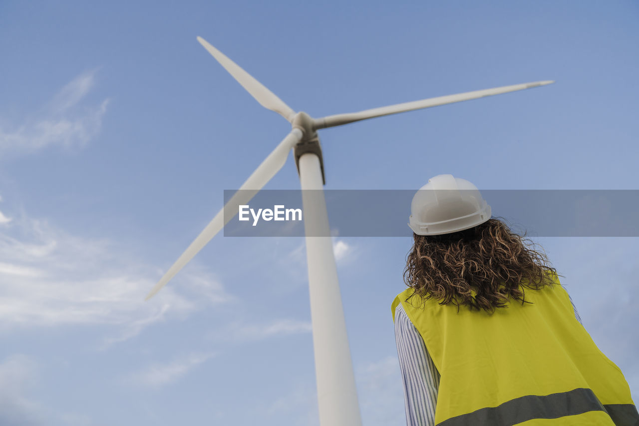 Engineer looking at rotor of tall wind turbine by blue sky