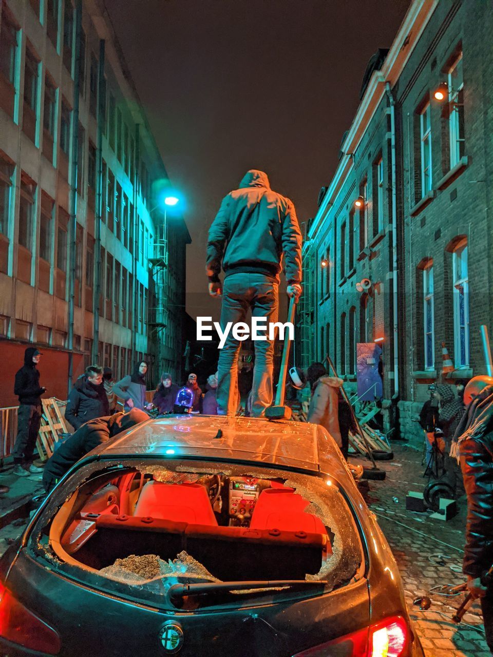 REAR VIEW OF PEOPLE STANDING ON ILLUMINATED STREET AMIDST BUILDINGS AT NIGHT