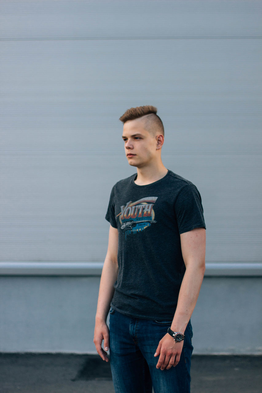 Young man looking away while standing against wall