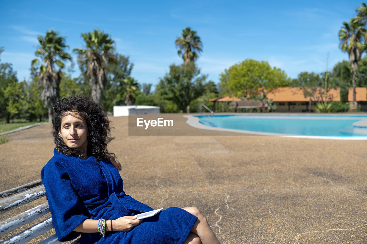 Woman relaxing sitting on a bench outdoors near swimming pool.