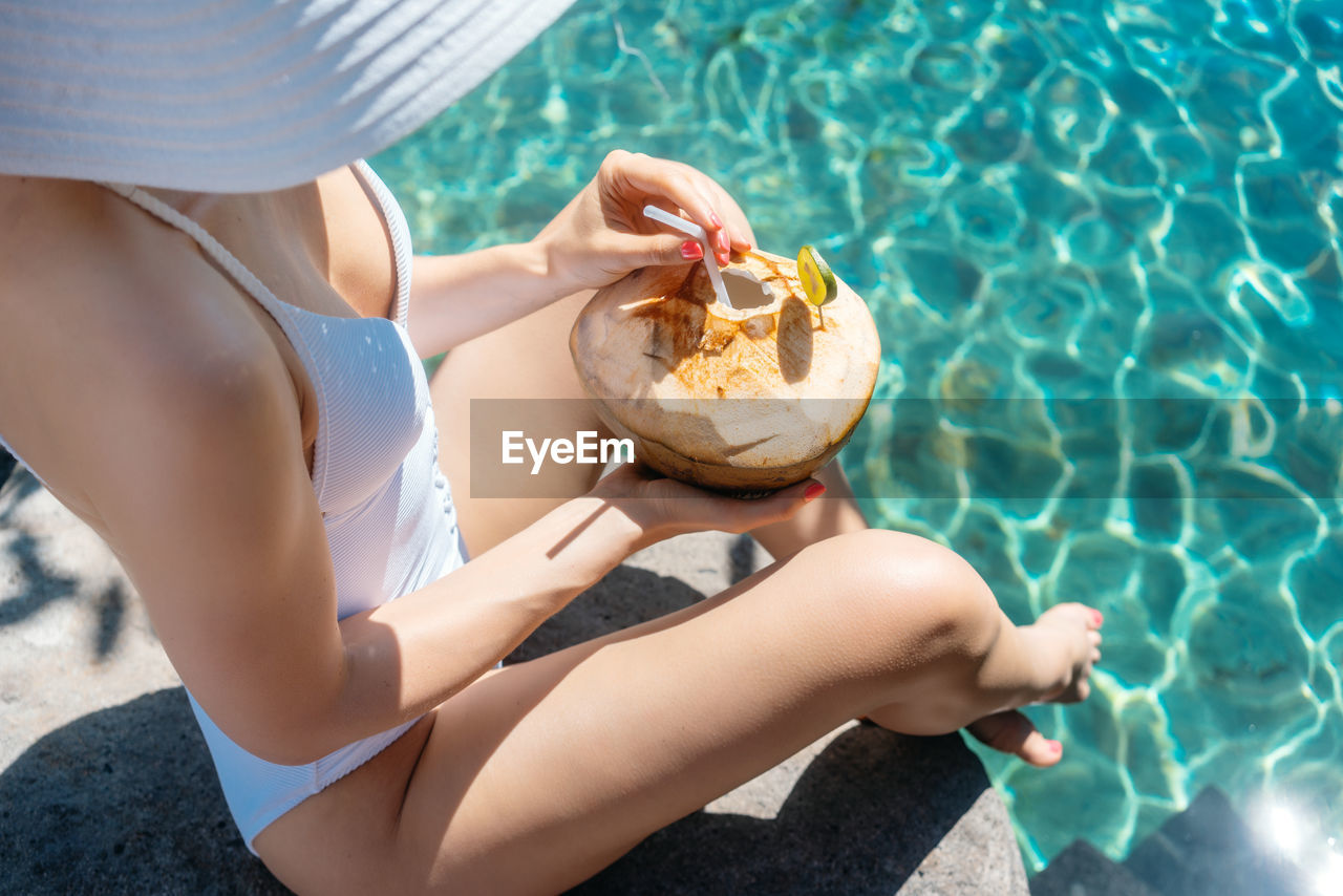 High angle view of woman holding coconut while sitting by swimming pool during sunny day
