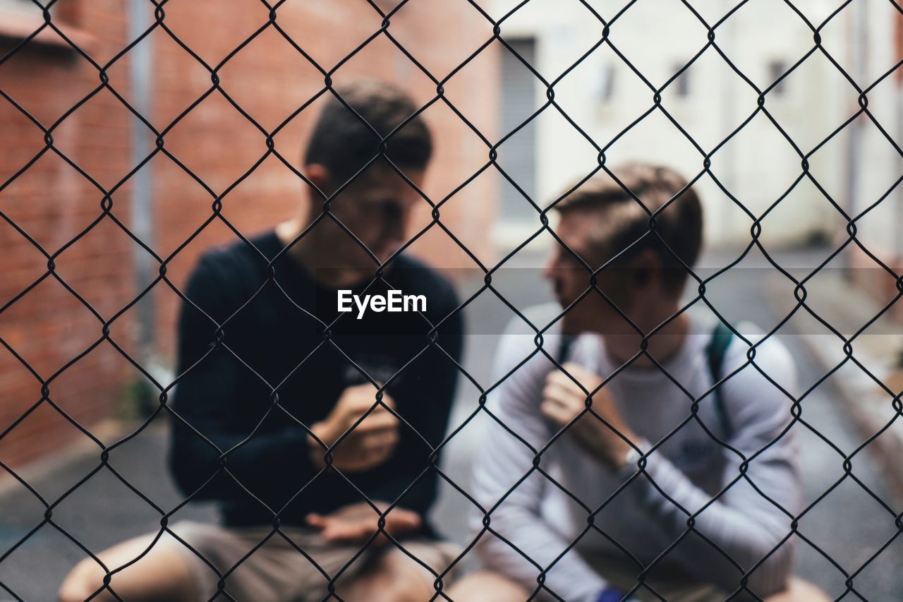 Male friends talking while sitting outdoors seen through chainlink fence