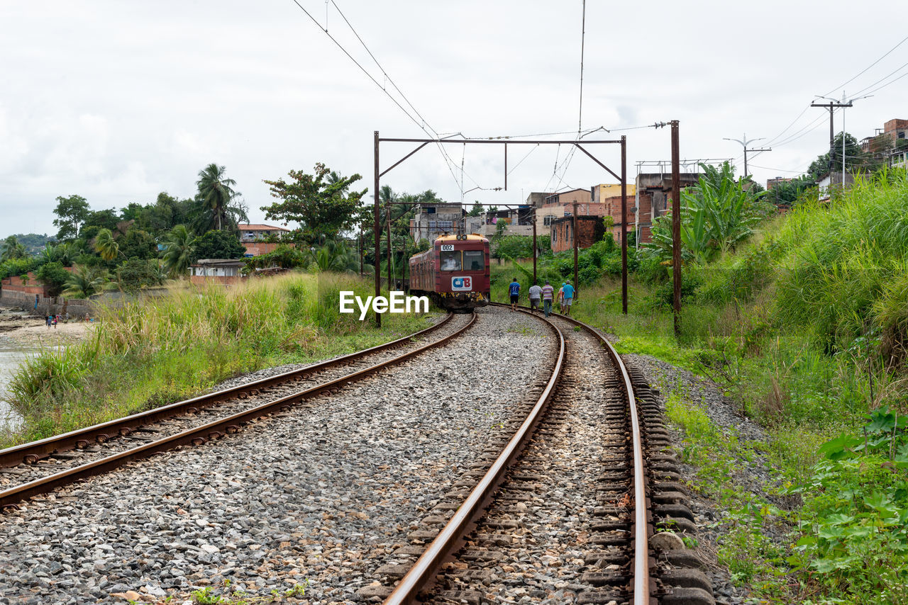 railroad track, rail transportation, track, transportation, mode of transportation, railway, sky, transport, cable, electricity, vehicle, public transportation, nature, train, plant, travel, power line, electricity pylon, architecture, rolling stock, cloud, no people, day, the way forward, technology, residential area, outdoors, tree, vanishing point, power supply, built structure, locomotive, diminishing perspective, environment