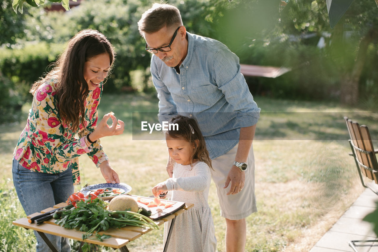 Girl cutting food while standing by mother and grandfather at table in backyard