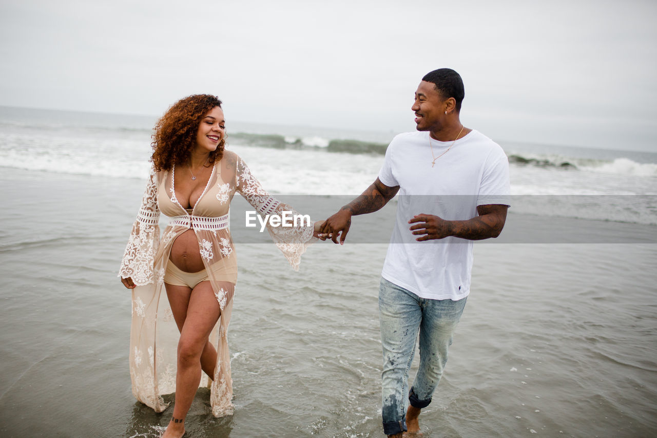 Mixed race couple running in water on beach, maternity