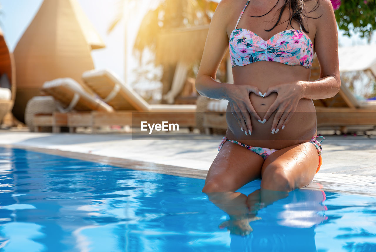 Low section of pregnant woman sitting heart shape on abdomen at poolside