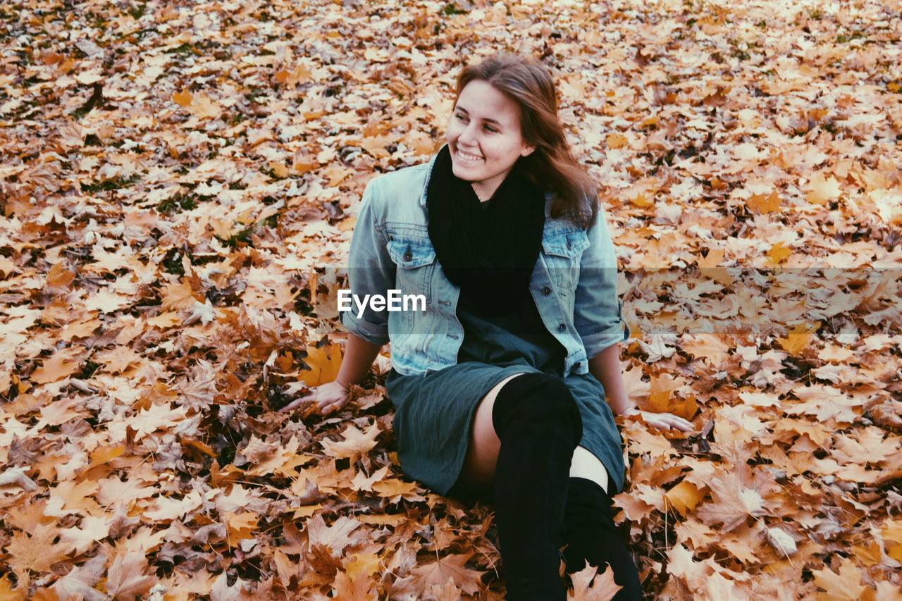 High angle view of smiling young woman sitting on field covered with autumn leaves