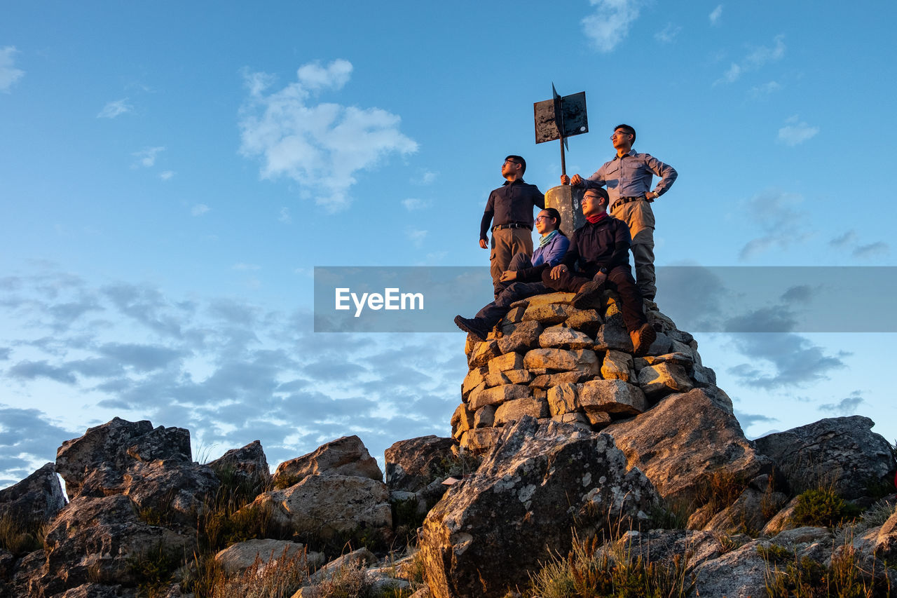 Four people standing on pile of rocks and trig point of mountain looking at the sunrise 