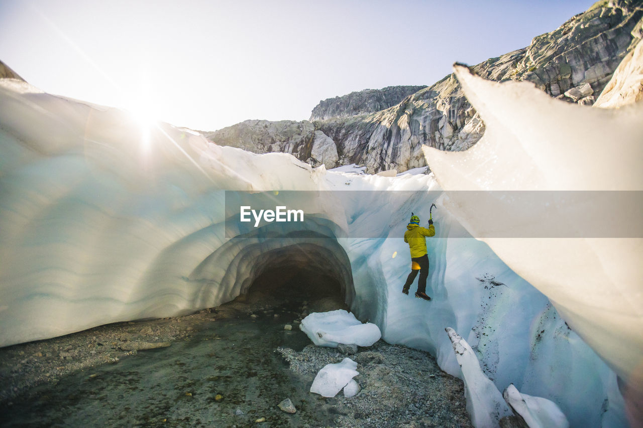 Rear view of man ice climbing outside of glacial cave.