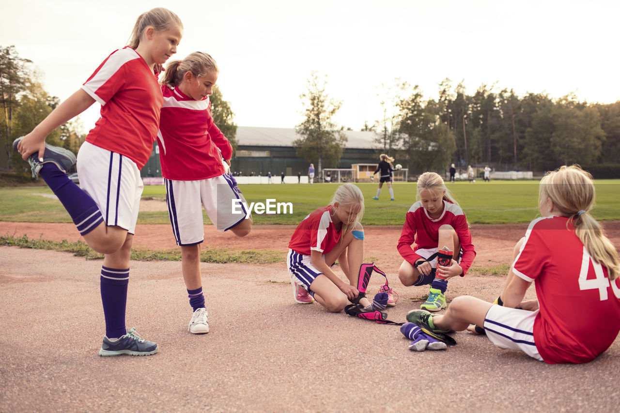 Girls stretching legs by friends wearing shin guards on field