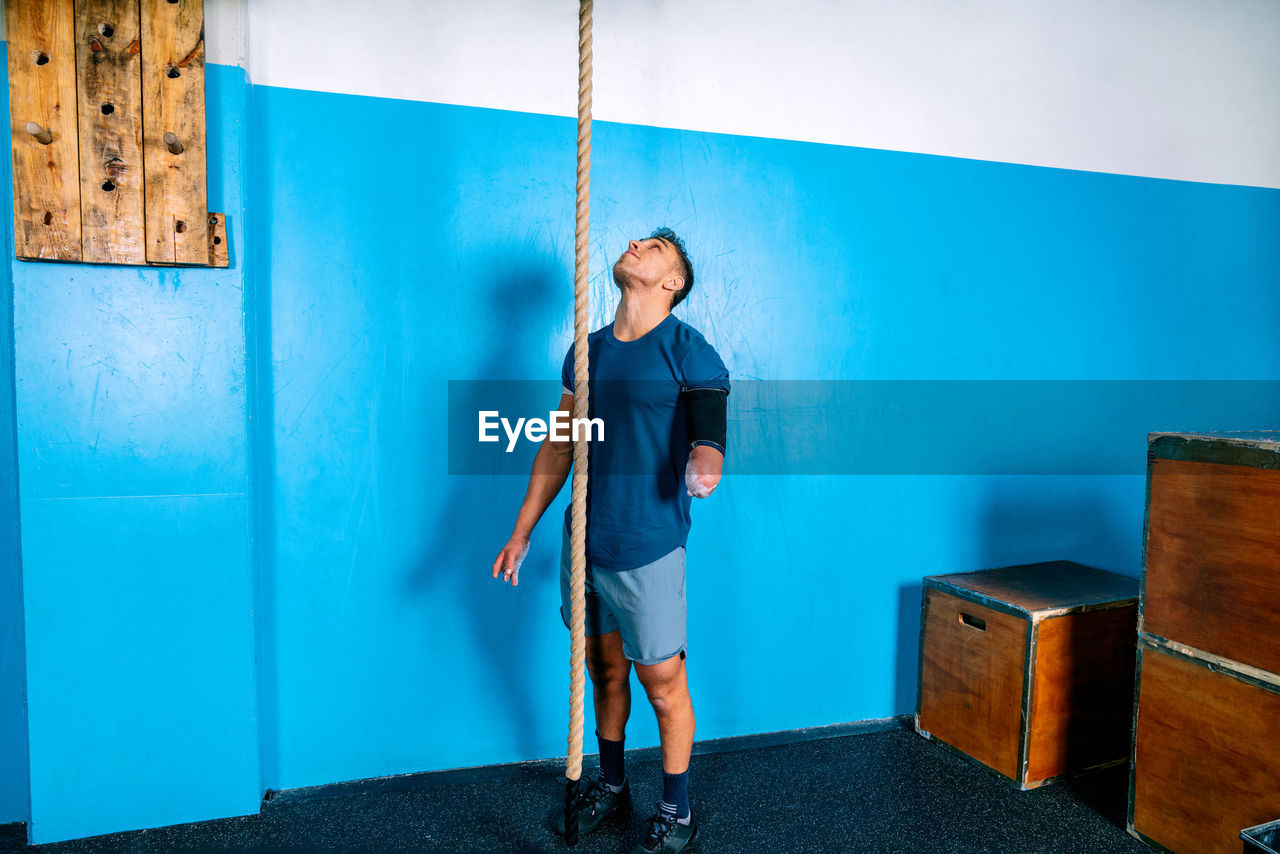 Disabled male athlete in sportswear looking up near workout rope and blue wall in gymnasium