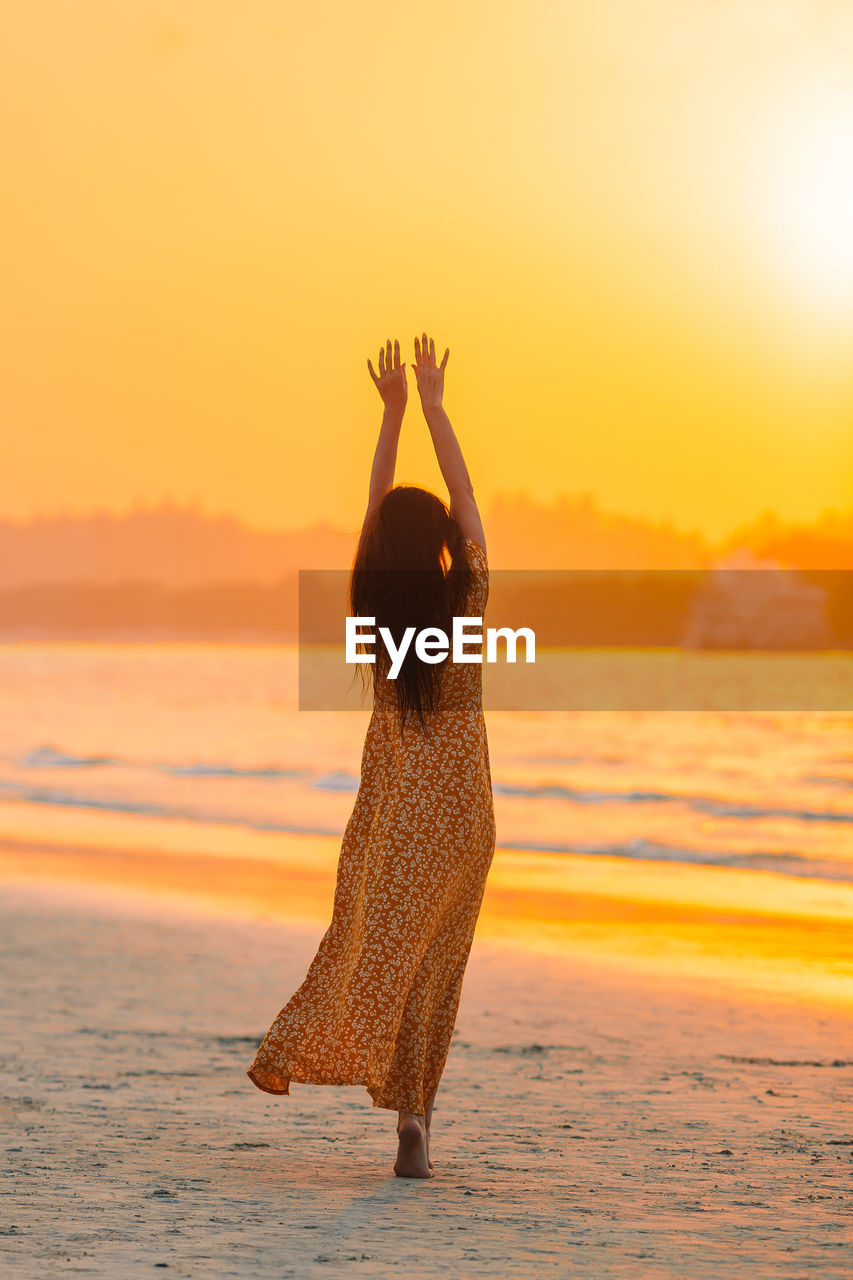 REAR VIEW OF WOMAN STANDING ON BEACH DURING SUNSET