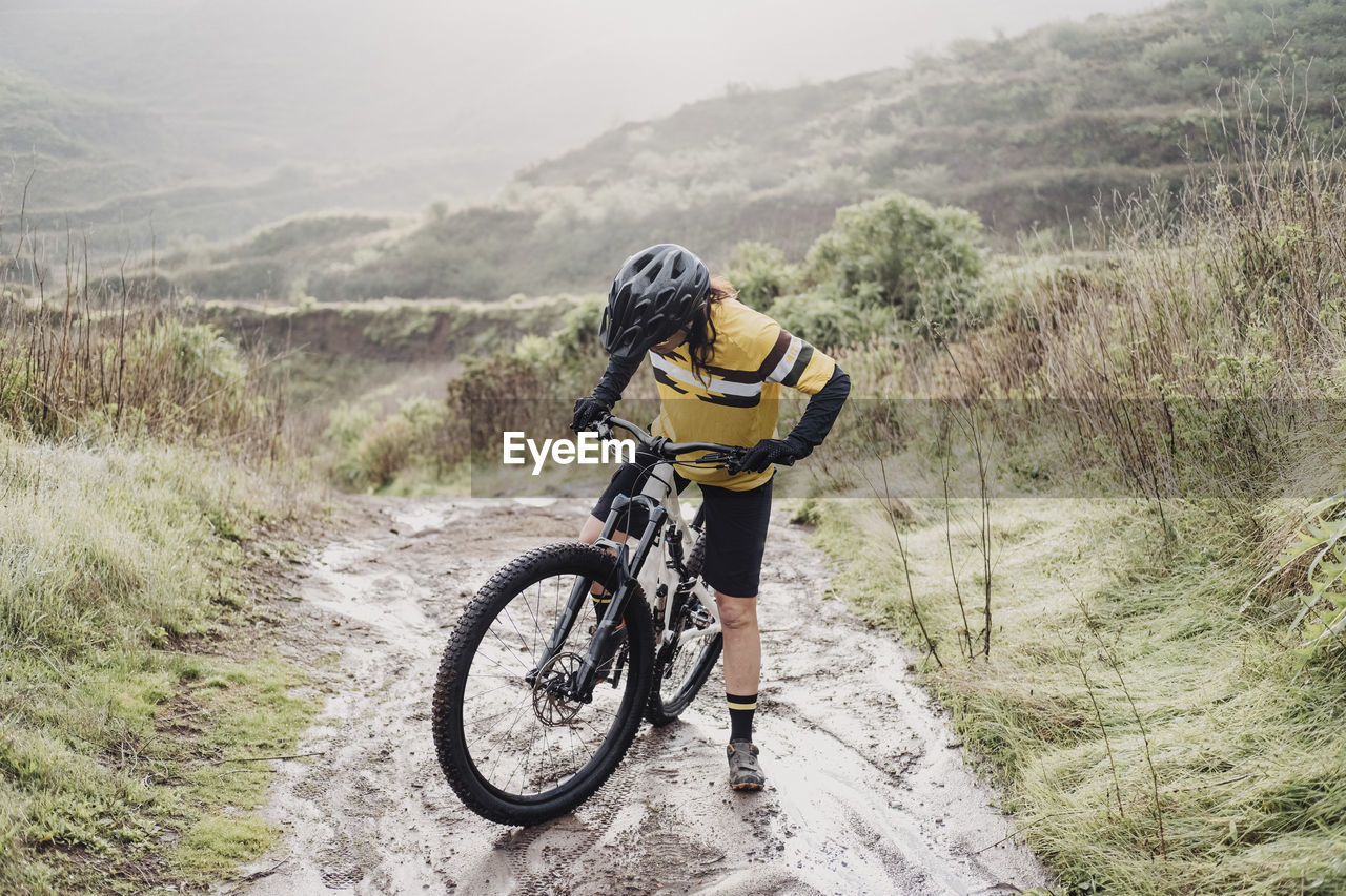 Full body of female cyclist on muddy mountain path on rainy day