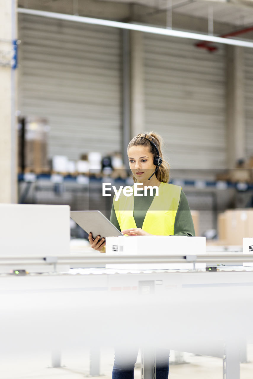 Worker wearing headset holding tablet pc examining box on conveyor belt in warehouse
