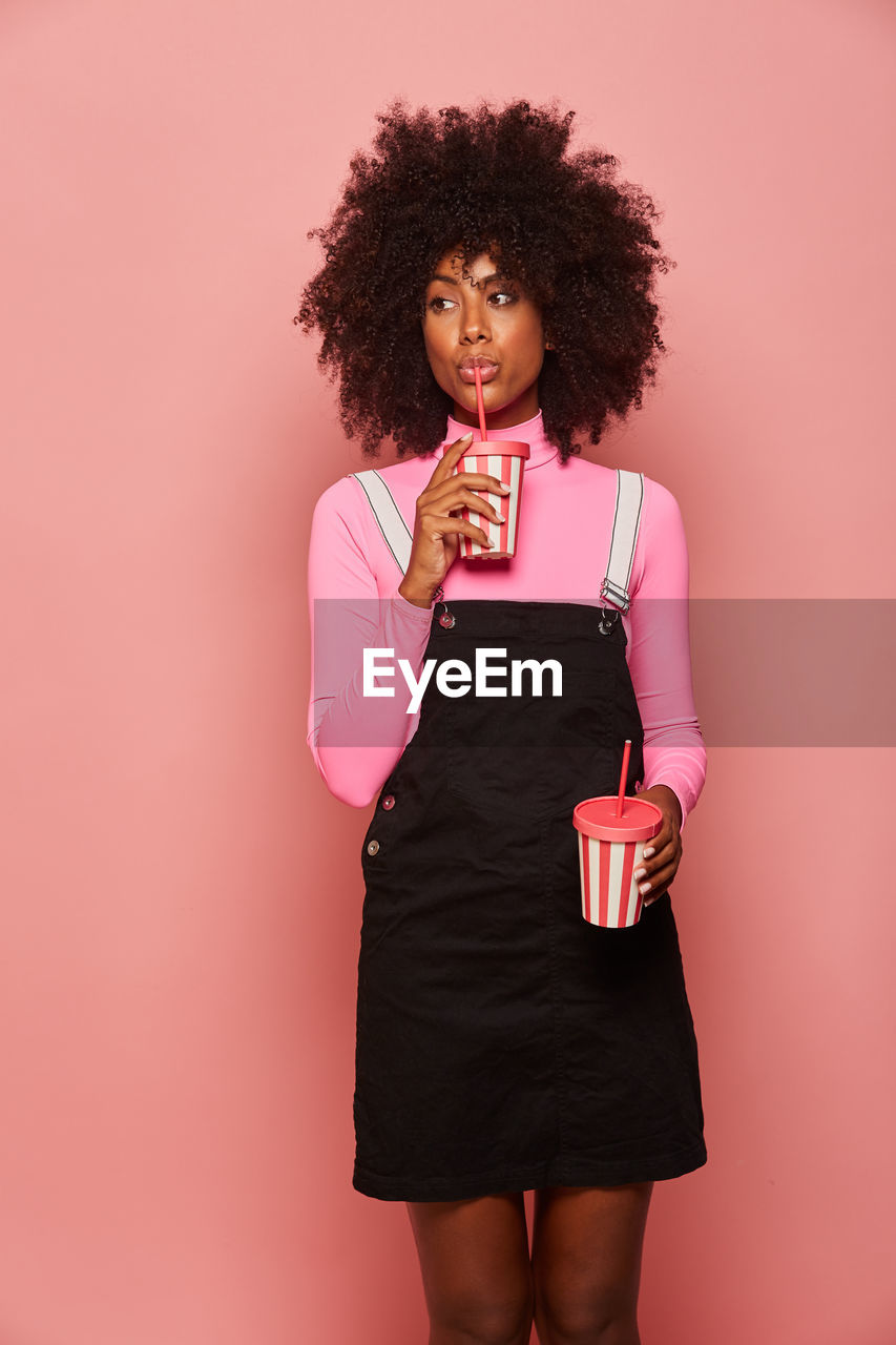 Happy black woman with disposable cup of drink standing against pink background