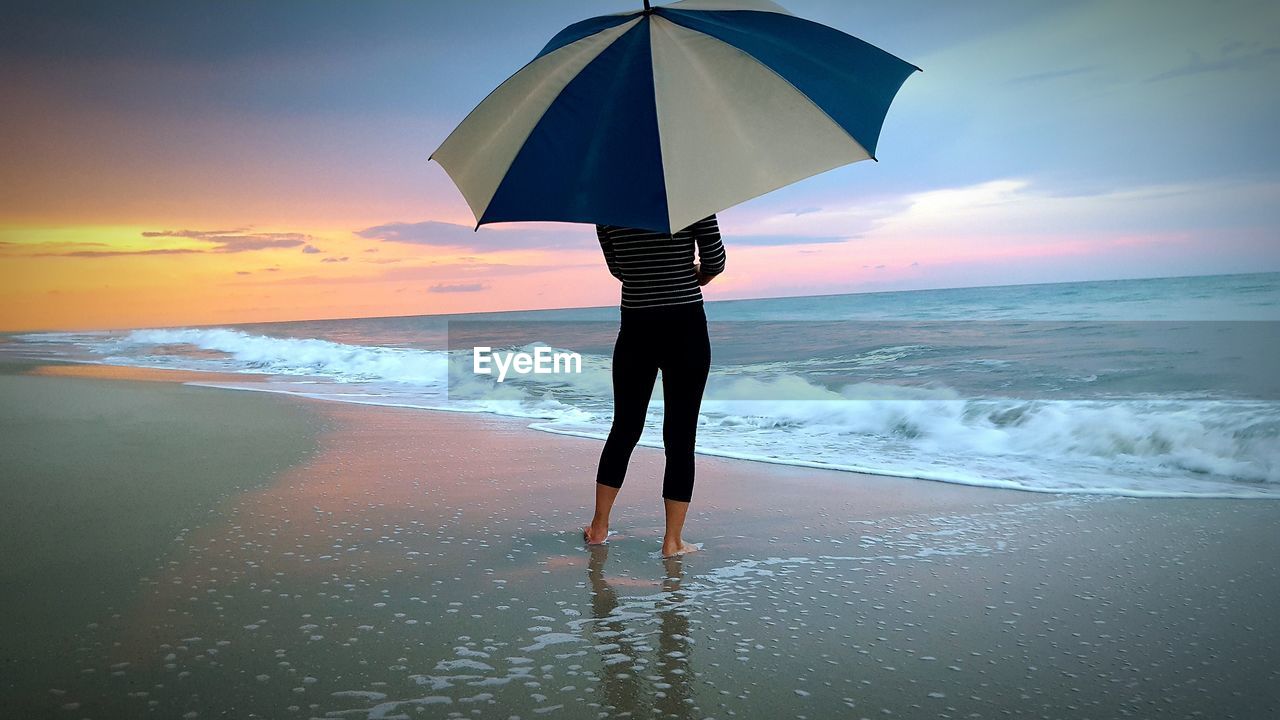 Woman with umbrella standing at beach against sky during sunset