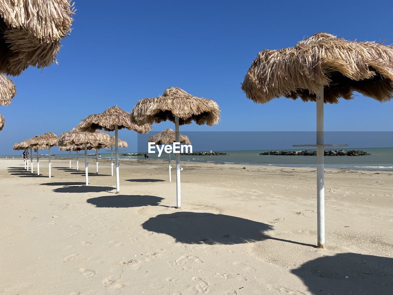 land, thatched roof, beach, water, sea, sky, nature, sand, sunlight, roof, ocean, parasol, blue, beauty in nature, sunny, coast, vacation, clear sky, tranquility, shore, beach umbrella, scenics - nature, day, shade, no people, shadow, summer, tranquil scene, umbrella, travel destinations, chair, sunshade, hut, tropical climate, holiday, outdoors, trip, relaxation, travel, idyllic, body of water, horizon