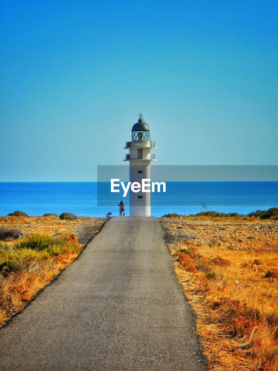 Mid distance of woman standing by lighthouse at beach against clear blue sky