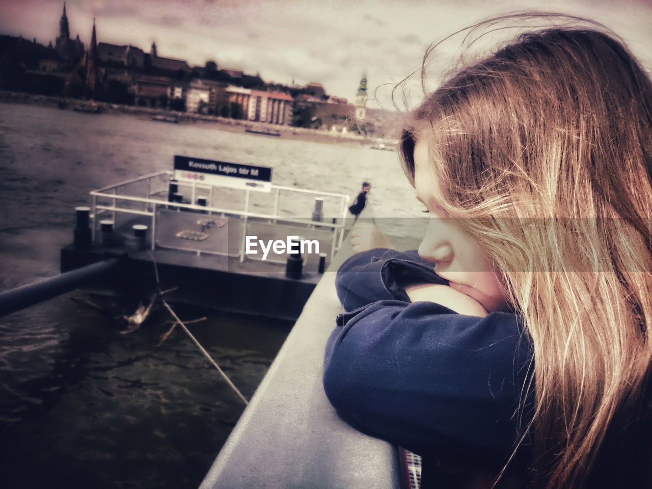 Side view of girl with blond hair leaning on railing by river in city