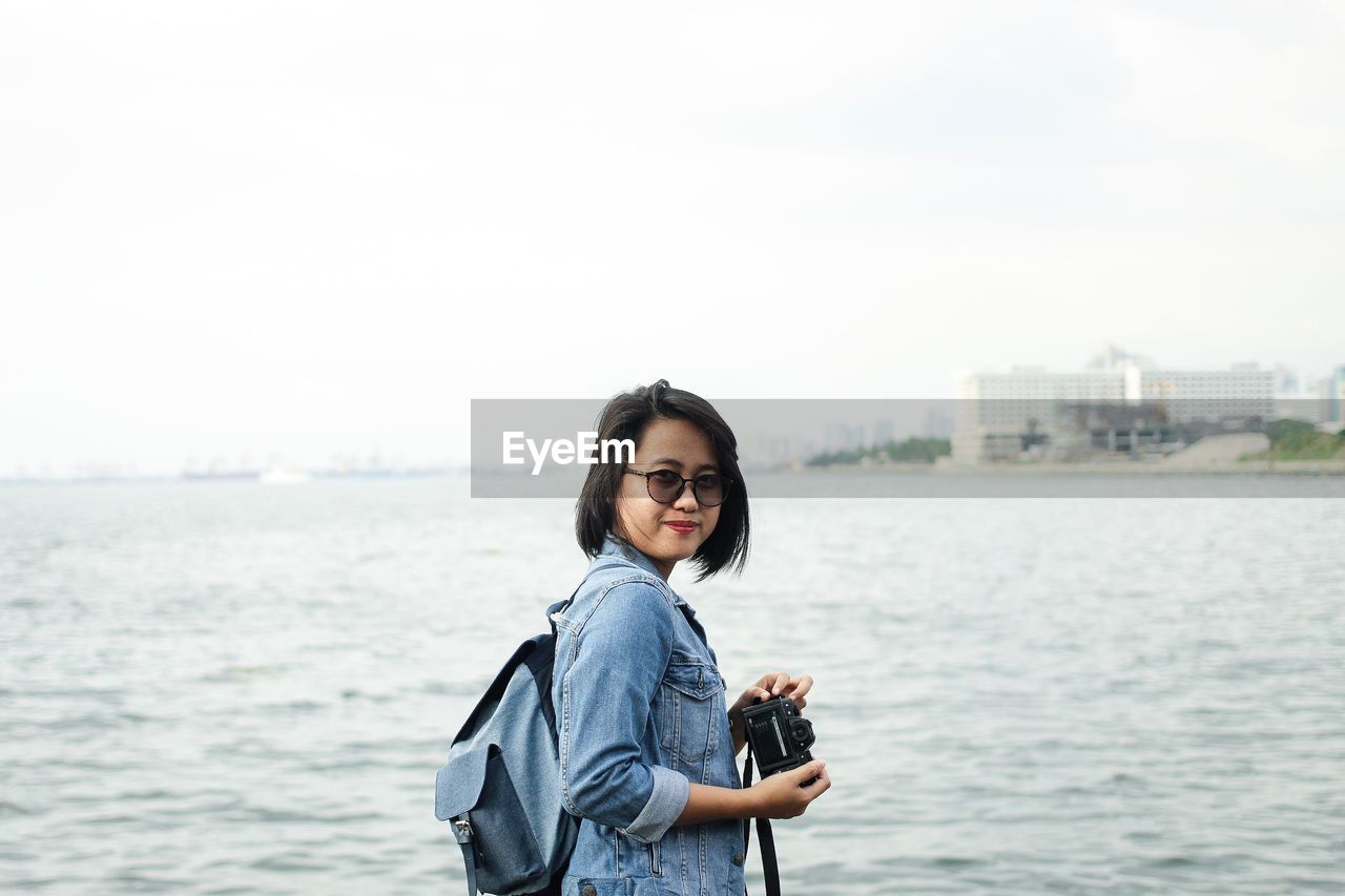 Portrait of smiling young woman standing by sea against sky