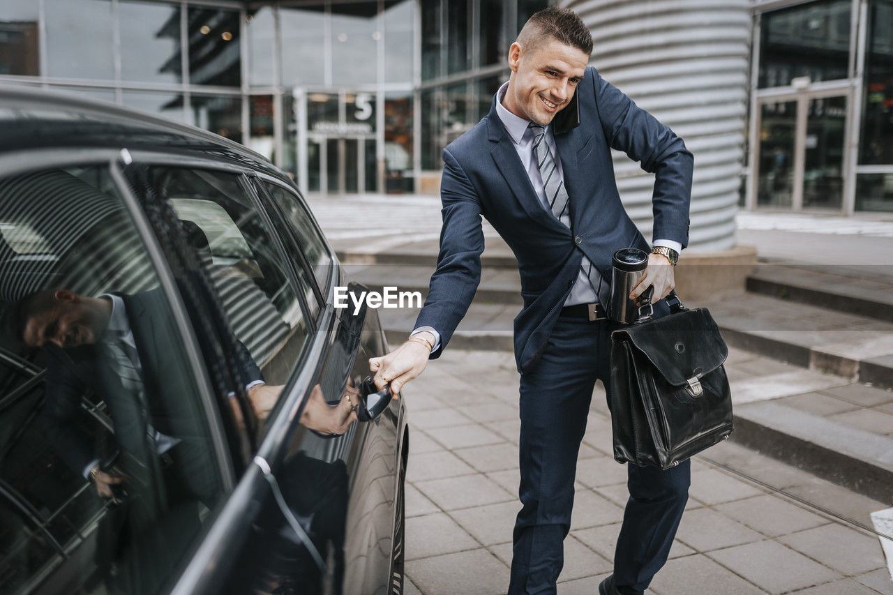 Businessman talking on smart phone while opening car door near office building