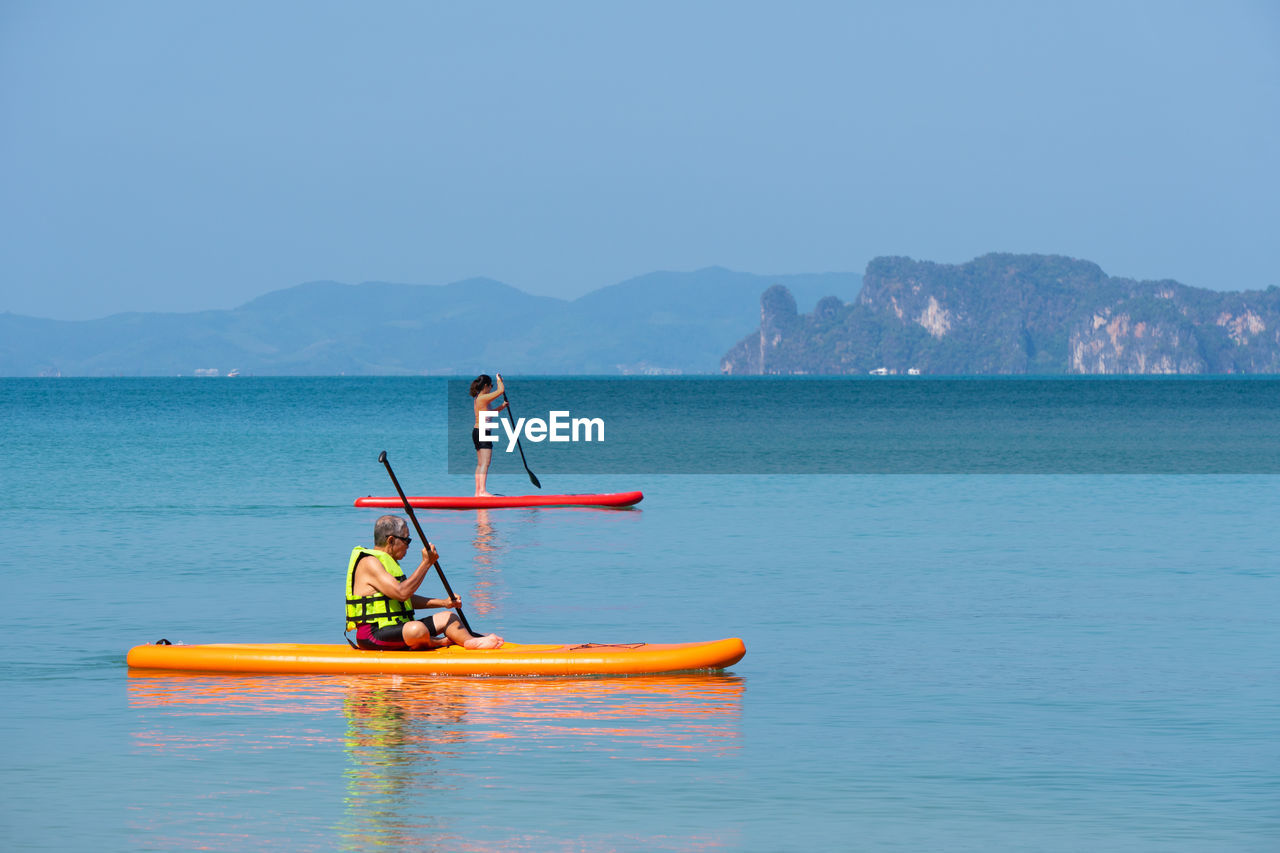 Asian senior father playing standup paddle board with young daughter at blue sea on summer vacation