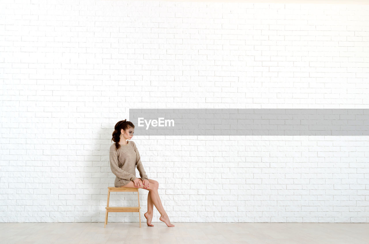 Full length of woman sitting on stool against wall