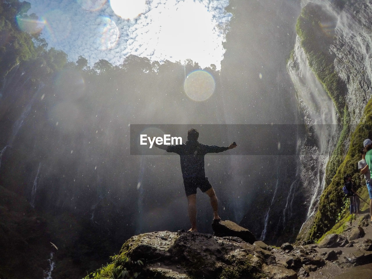 Full length of male hiker with arms outstretched standing against waterfall