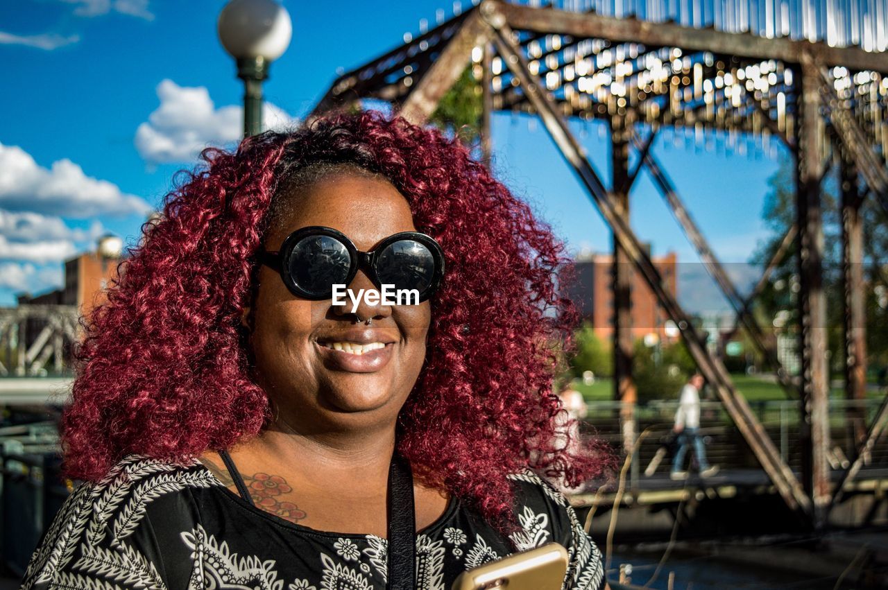 Portrait of woman with curly maroon hair while wearing sunglasses against bridge
