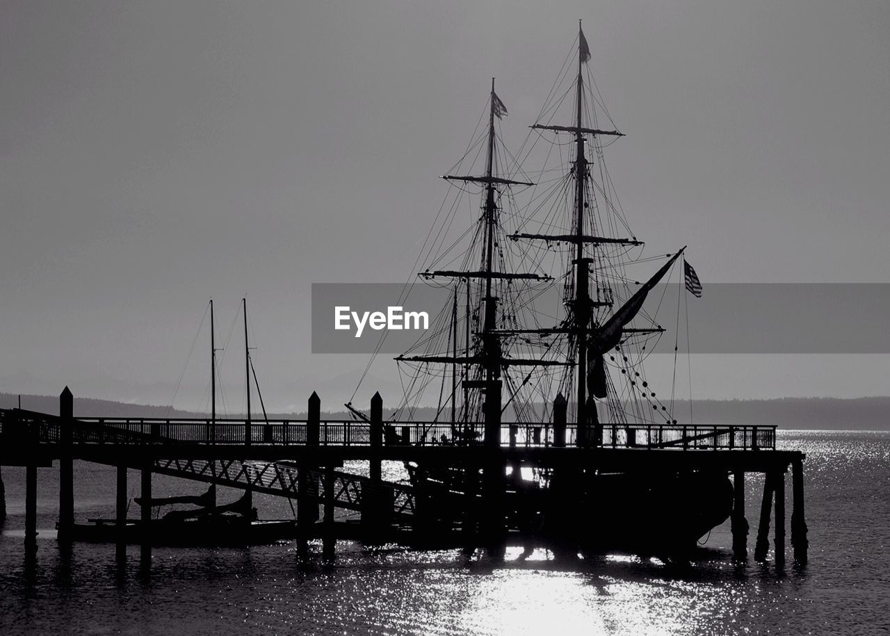 Silhouette sailing ship moored on jetty in sea water against sky