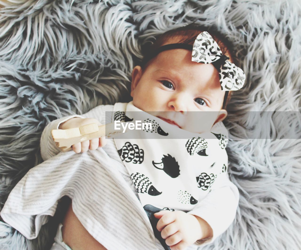 Portrait of cute baby lying on bed at home. monochrome colours, bow, hedgehogs bib, wooden teether.