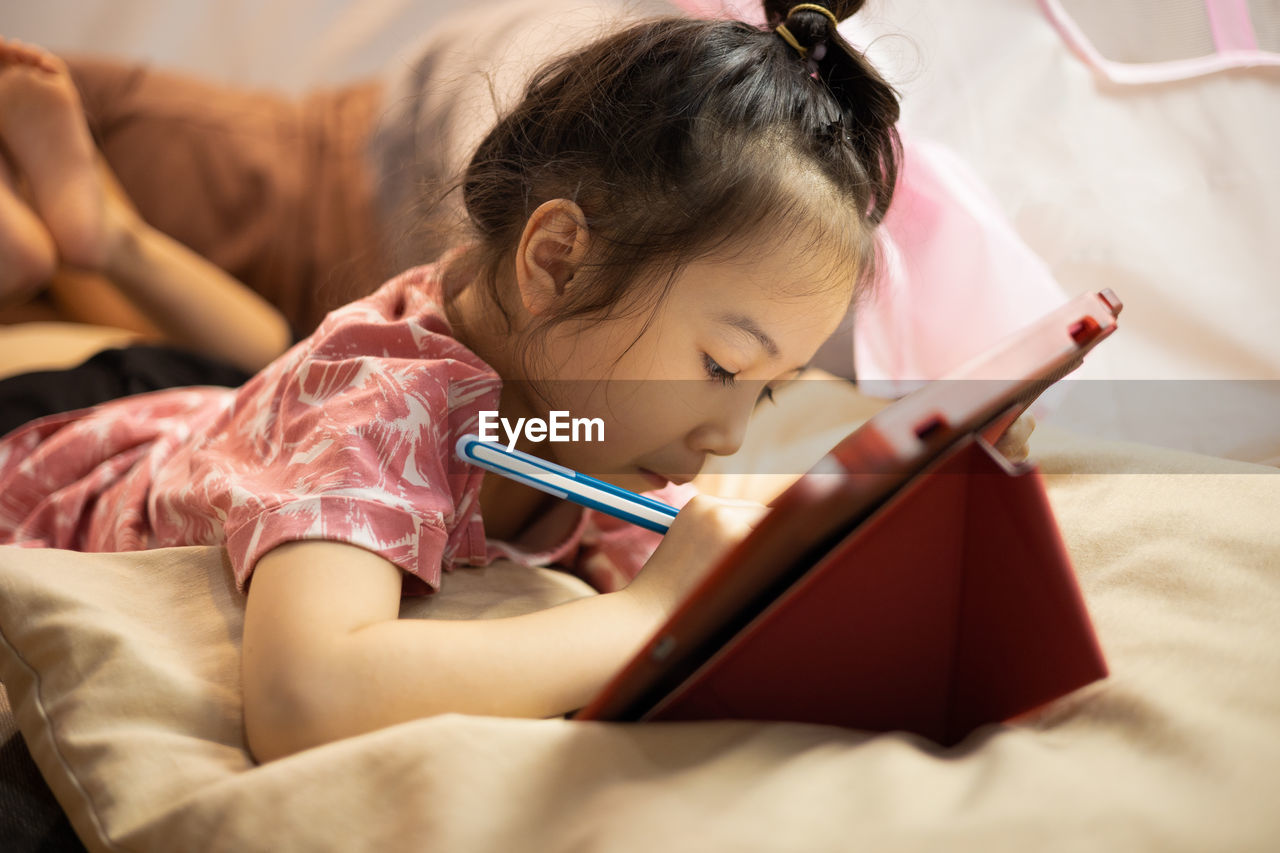 Asian adorable kid girl is lying on the floor in living room at home and writing on tablet 