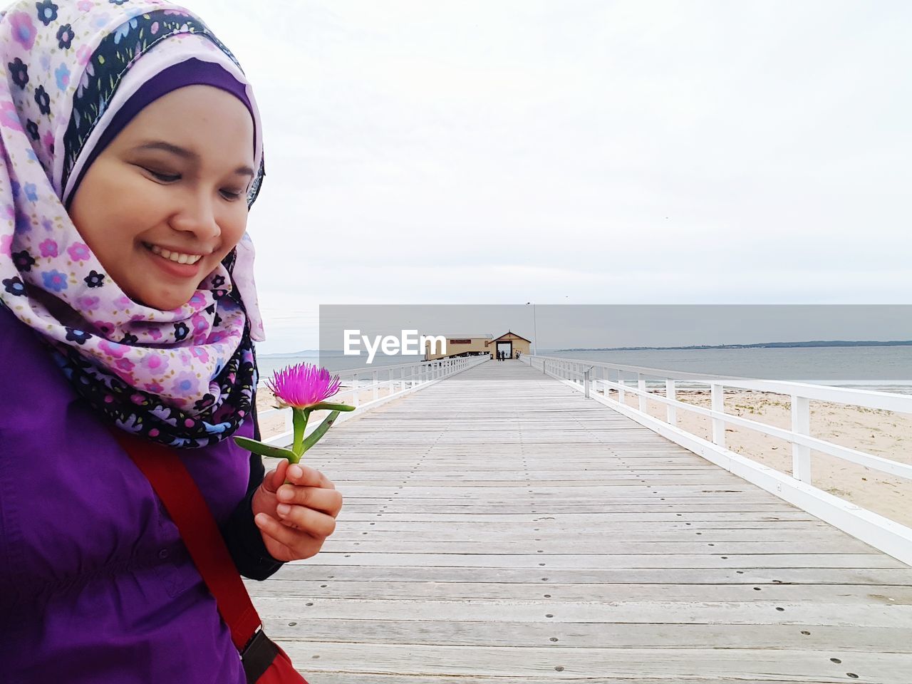 Smiling woman in hijab holding flower on pier at sea against sky