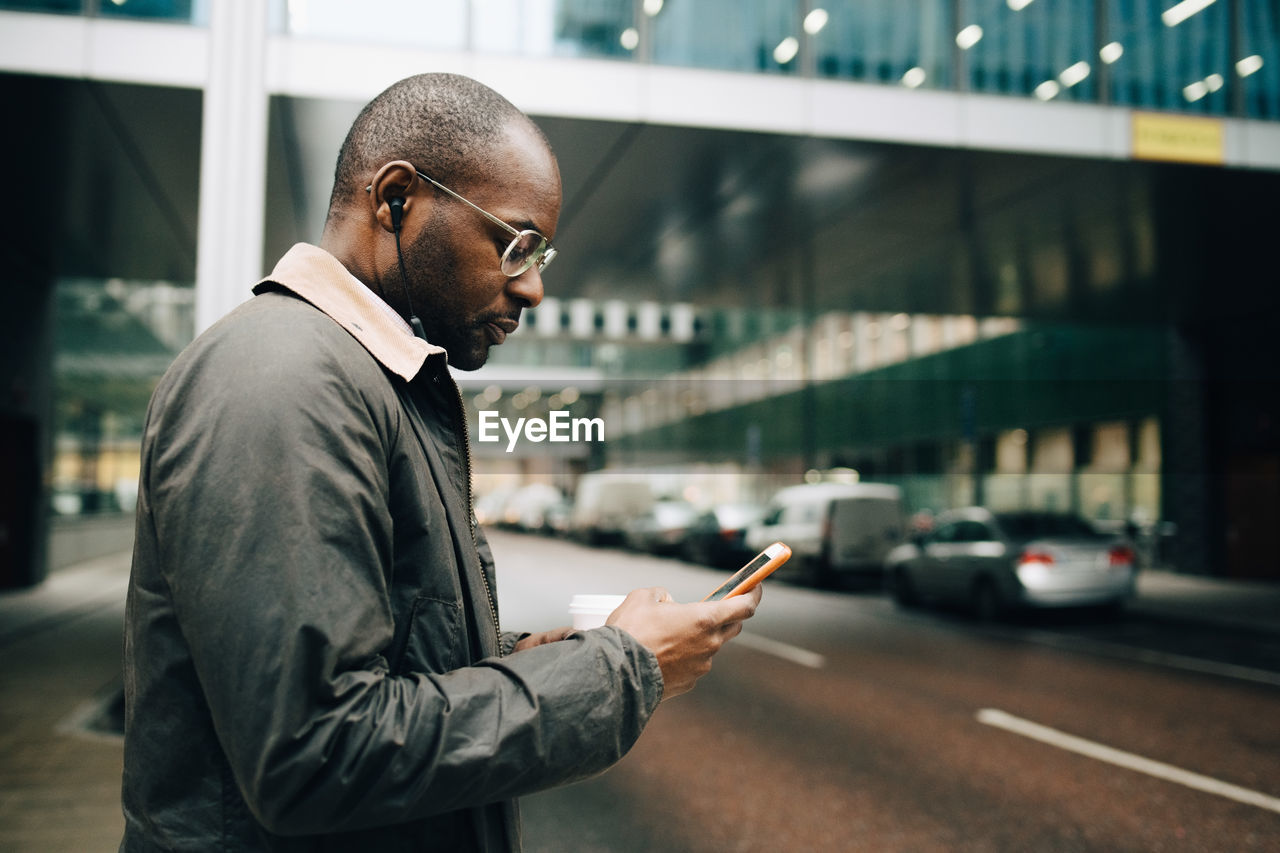 Side view of businessman using mobile phone while standing on street against building in city