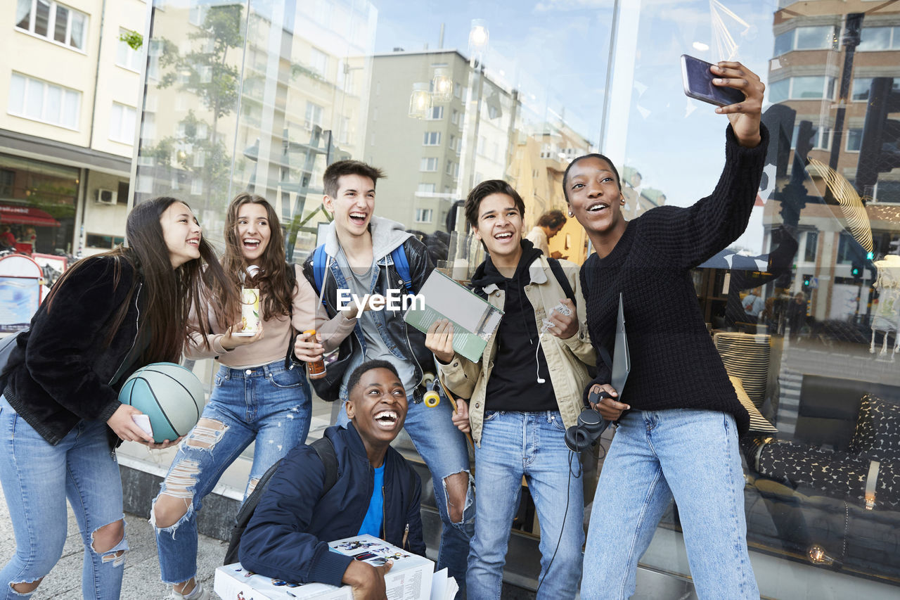 Cheerful teenage girl taking selfie with friends on footpath by store in city