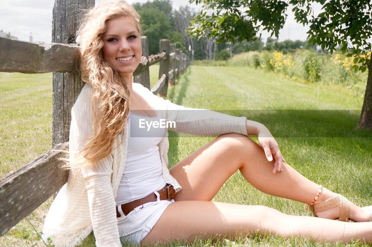 Portrait of beautiful woman sitting on grassy field against wooden fence
