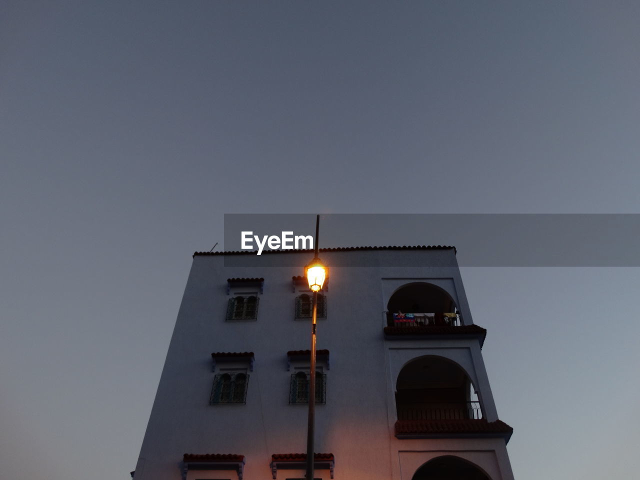 Low angle view of illuminated street light by building against sky at dusk
